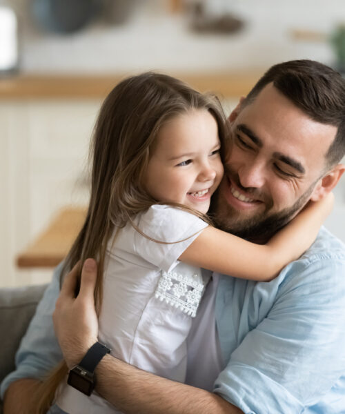 Close up smiling loving young father hugging adorable little daughter, enjoying tender moment, spending weekend together, sitting on cozy couch at home, good family relationship between dad and child