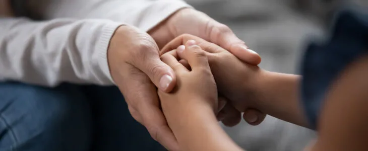 Close up compassionate young foster parent holding hands of little kid girl, giving psychological help, supporting at home. Sincere different generations family sharing secrets or making peace.
